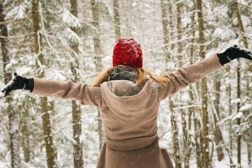girl standing in the snow with gloves, tan coat and red hat on